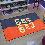 Pop art ve modern grafik estetiğinde tasarlanmış dekoratif halı, koyu mavi zemin üzerinde merkezde büyük ve kalın tipografi ile yazılmış “LET’S STAY LOUD” metni içerir. Turuncu dalga formunda renk bloğu yazının arkasında güçlü kontrast oluşturur ve minimal tipografi odaklı grafik tasarım kompozisyonu sunar.